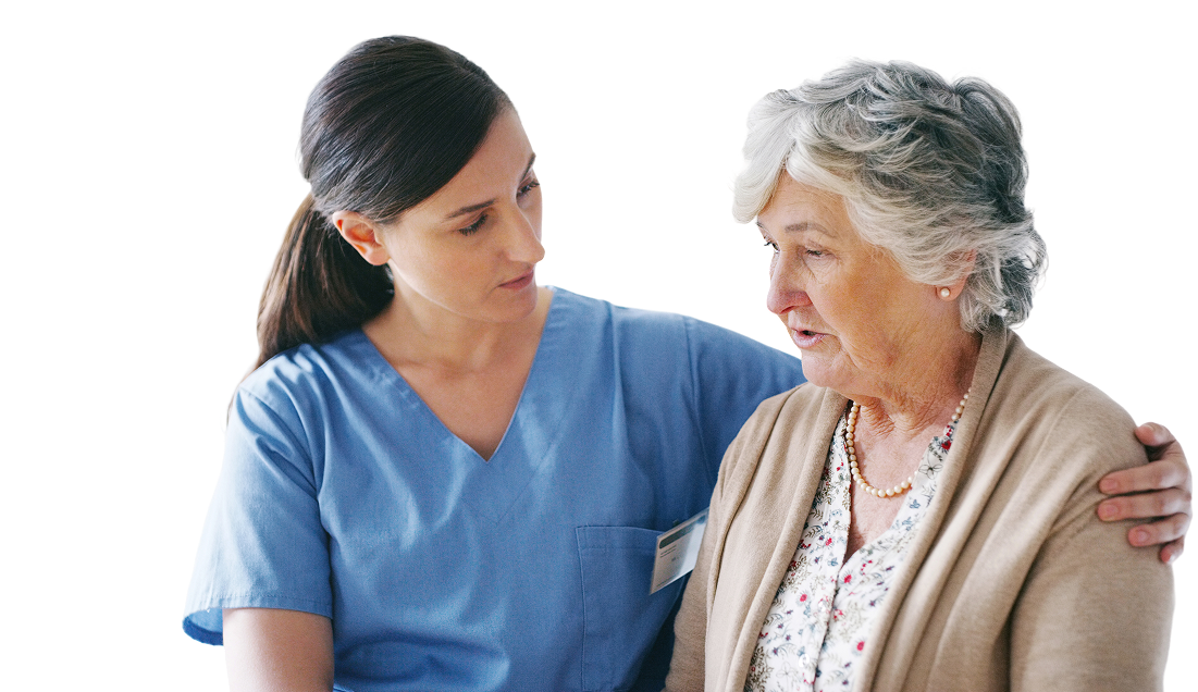 A healthcare worker in blue scrubs with a name tag is sitting beside an elderly person with gray hair, wearing a beige cardigan over a floral blouse. The healthcare worker has one arm around the elderly person's shoulder, providing support and comfort.
